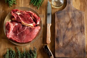 Top view of chef knife next to big chunks of red meat and green vegetables on wooden table.